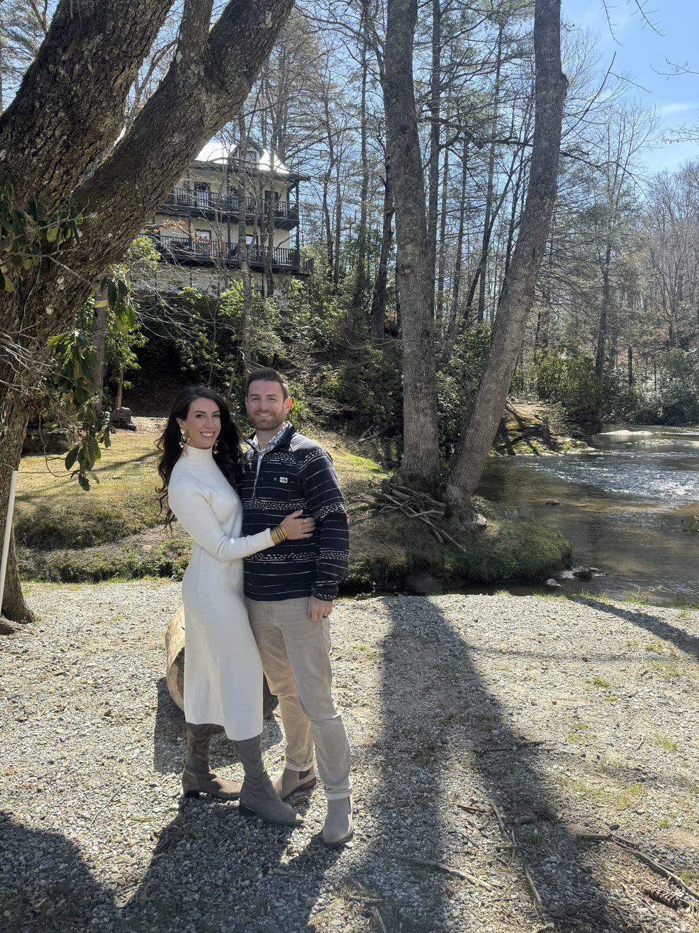 Two people standing on the gravel riverbank in winter sunlight, with the retreat home's porches visible on the hillside behind them.