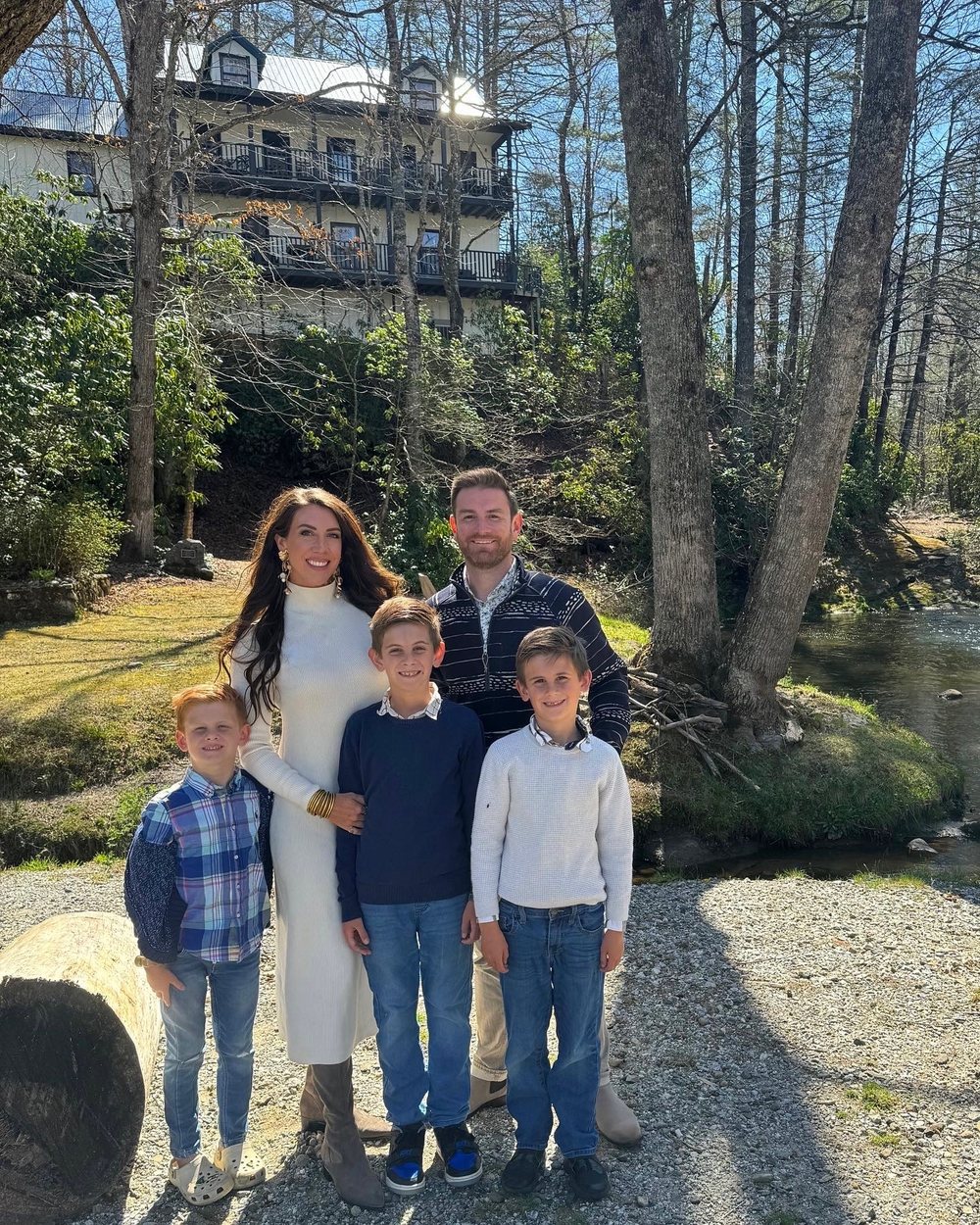 A family of five standing together on the gravel riverbank in spring sunshine, the river beside them and the retreat home's porches on the hillside behind.